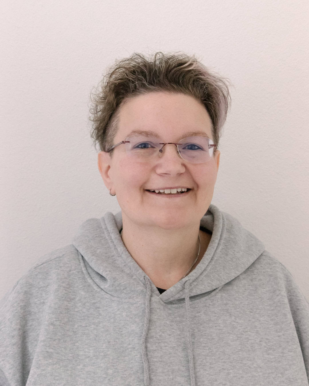 1. Frau mit kurzer, lockiger Frisur und Brille, lächelnd, trägt grauen Hoodie.